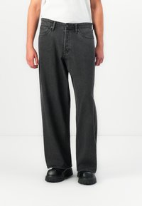 Mann som har på seg løse svarte denimjeans, svarte sko med tykk såle, og en hvit skjorte, står mot en enkel hvit bakgrunn.