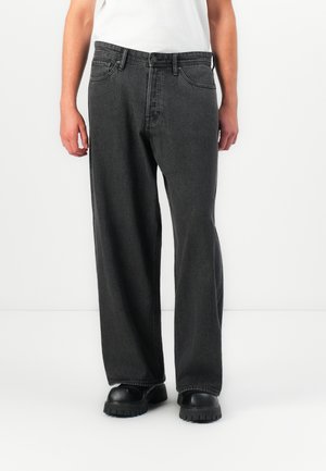 Hombre que lleva puestos unos jeans de mezclilla negros holgados, zapatos negros de suela gruesa y una camisa blanca, de pie contra un fondo blanco liso.
