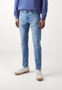 Mannen bär ljusblå slim-fit jeans, vita sneakers med gummisulor och en lila sweatshirt, stående mot en plain vit bakgrund.