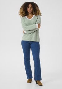 Maglione a maglia verde chiaro con scollo a V, indossato sopra una camicia bianca a maniche lunghe. Abbinato a jeans a zampa d'elefante blu e stivali con tacco marroni.