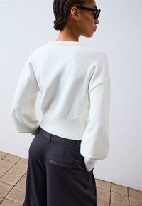 Biely pletený sveter s rebrovaným lemom a prehodenými ramenami, v kombinácii s tmavošedými upravenými nohavicami s náprsným vreckom.