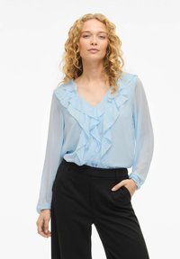 Lyseblå bluse med lagdelte volanger, V-hals og lange ermer. Kombinert med svarte, høytlivete bukser for et sofistikert utseende.