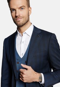 Granatowy garnitur w kratkę, z białą koszulą z kołnierzykiem i kamizelką w kratę. Guziki na kamizelce są brązowe. Widoczny tatuaż na nadgarstku.