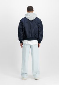 Hombre de espaldas vistiendo una chaqueta bomber azul marino sobre una sudadera gris, una camiseta blanca, unos pantalones vaqueros de pierna ancha en azul claro, y zapatillas crema.