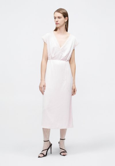 Γυναίκα με ροζ λιλά μίντι φόρεμα με δαντέλα στο τελείωμα, συνδυασμένο με λευκές καλτσοπότες με σχέδια και μαύρα ψηλοτάκουνα σανδάλια, στέκεται σε λευκό φόντο.