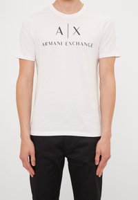 Witte T-shirt met korte mouwen en een ronde hals. Voorzien van een zwart logo met de tekst "A|X ARMANI EXCHANGE" op de voorkant. Zachte katoenstof.