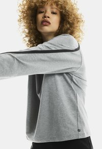 Young person with curly hair wearing a light gray long-sleeve shirt with black stripe detail stretching arm forward.