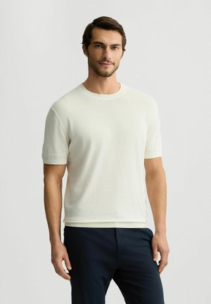 Kurzärmliges T-Shirt mit Rundhalsausschnitt in hellem Creme, aus weichem Stoff mit subtiler Struktur und gerippten Ärmeln, kombiniert mit dunklen Hosen.