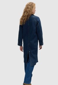 Długi płaszcz dżinsowy w ciemnoniebieskim kolorze z rozcięciem z tyłu, długimi rękawami i zapięciem na zatrzask. Stylizowany na niebieskich dżinsach, podkreślający mocny materiał i detale.