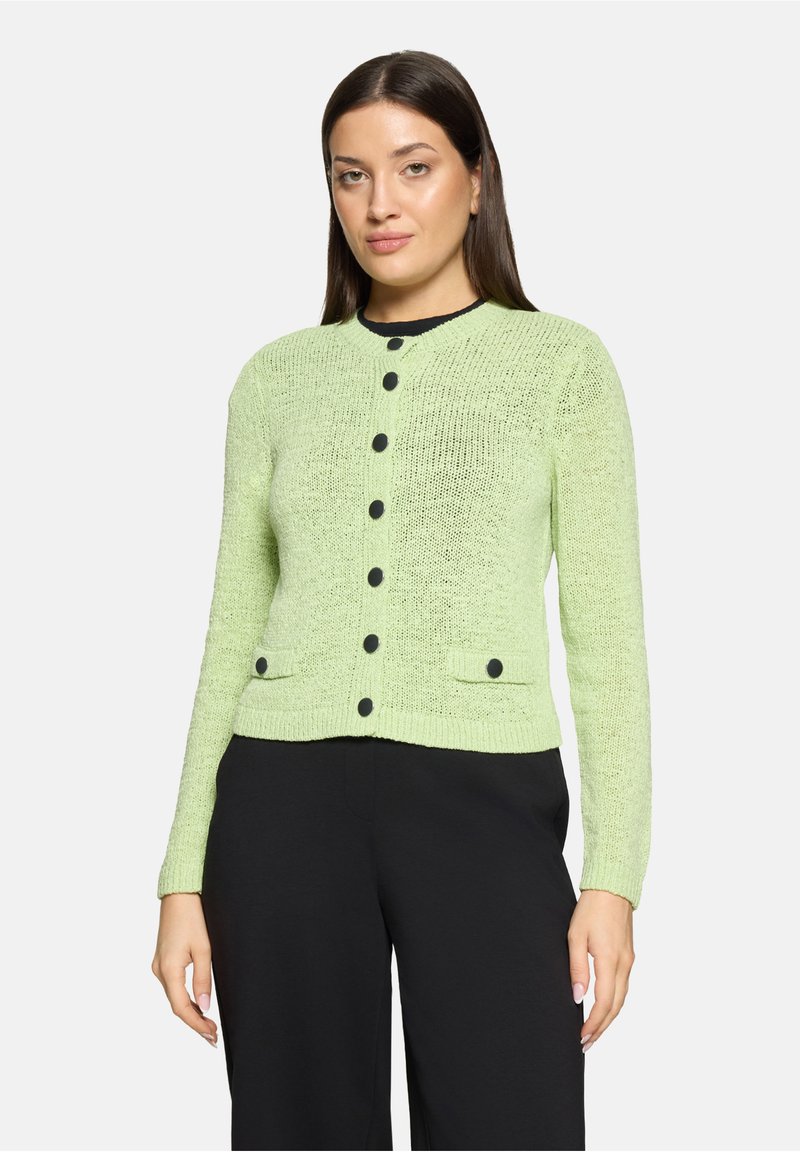 Kobieta ubrana w jasnozielony dziergany kardigan z czarnymi guzikami, nosząca czarne spodnie, stoi na tle jednolitego białego tła.