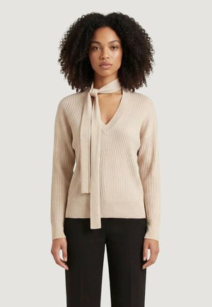 Donna con capelli ricci che indossa un maglione beige a costine con scollo a V e un dettaglio di cravatta lunga, abbinato a pantaloni neri, in piedi davanti a uno sfondo neutro.