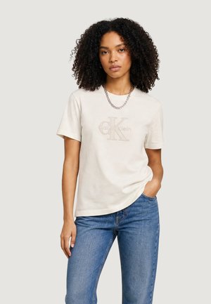 Kobieta o kręconych włosach nosi jasnobeżowy T-shirt Calvin Klein, niebieskie dżinsy i srebrny naszyjnik z łańcuszkiem, stojąc na jednolitym tle.