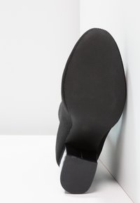 Bottine noire à talon, positionnée semelle vers le haut contre un mur d'angle blanc et le sol, mettant en valeur la semelle lisse et le talon texturé.