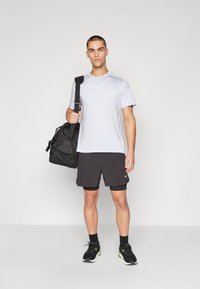 Conjunto deportivo que incluye una camiseta gris claro, pantalones cortos negros y zapatillas negras. El modelo sostiene una bolsa de gimnasio negra con una correa.
