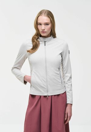 Hosszú szőke hajú nő fehér, testhezálló, cipzáras dzsekit és légies mályvaszínű szoknyát visel, egyszínű világos háttér előtt áll.