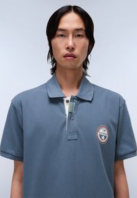 Modrá polo triko s texturovanou látkou, tříknoflíkovou légou a logem na levé hrudi. Krátké rukávy a design límce.