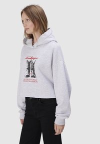 Kobieta w jasnoszarej krótkiej bluzie z kapturem i czarnych dżinsach, z grafiką kowbojskich butów i czerwonym napisem na przodzie.