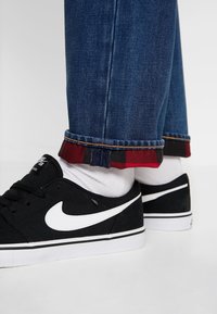 Sneakers in tela nera con logo Nike bianco e suole bianche, abbinati a jeans blu scuro con risvolto in plaid rosso e nero.