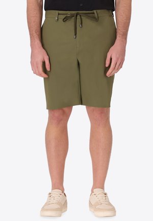 Uomo che indossa pantaloncini di colore verde oliva fino al ginocchio con coulisse in vita e scarpe da ginnastica beige con lacci, in piedi davanti a uno sfondo semplice.