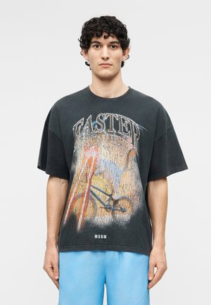 Νέος άνδρας με σγουρά μαλλιά που φοράει μαύρο μπλουζάκι με γραφικά με ένα ποδήλατο και το κείμενο «WASTER OCEAN LIFE», σε συνδυασμό με ανοιχτόχρωμα μπλε σορτς.