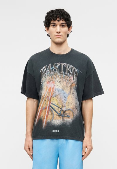 Jeune homme aux cheveux bouclés portant un t-shirt noir graphique représentant un vélo avec le texte "WASTER OCEAN LIFE", associé à un short bleu clair.