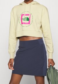 Felpa cropped beige con cappuccio e stampa del logo verde e rosa; abbinata a una gonna pantalone blu navy con tasca laterale e texture liscia.