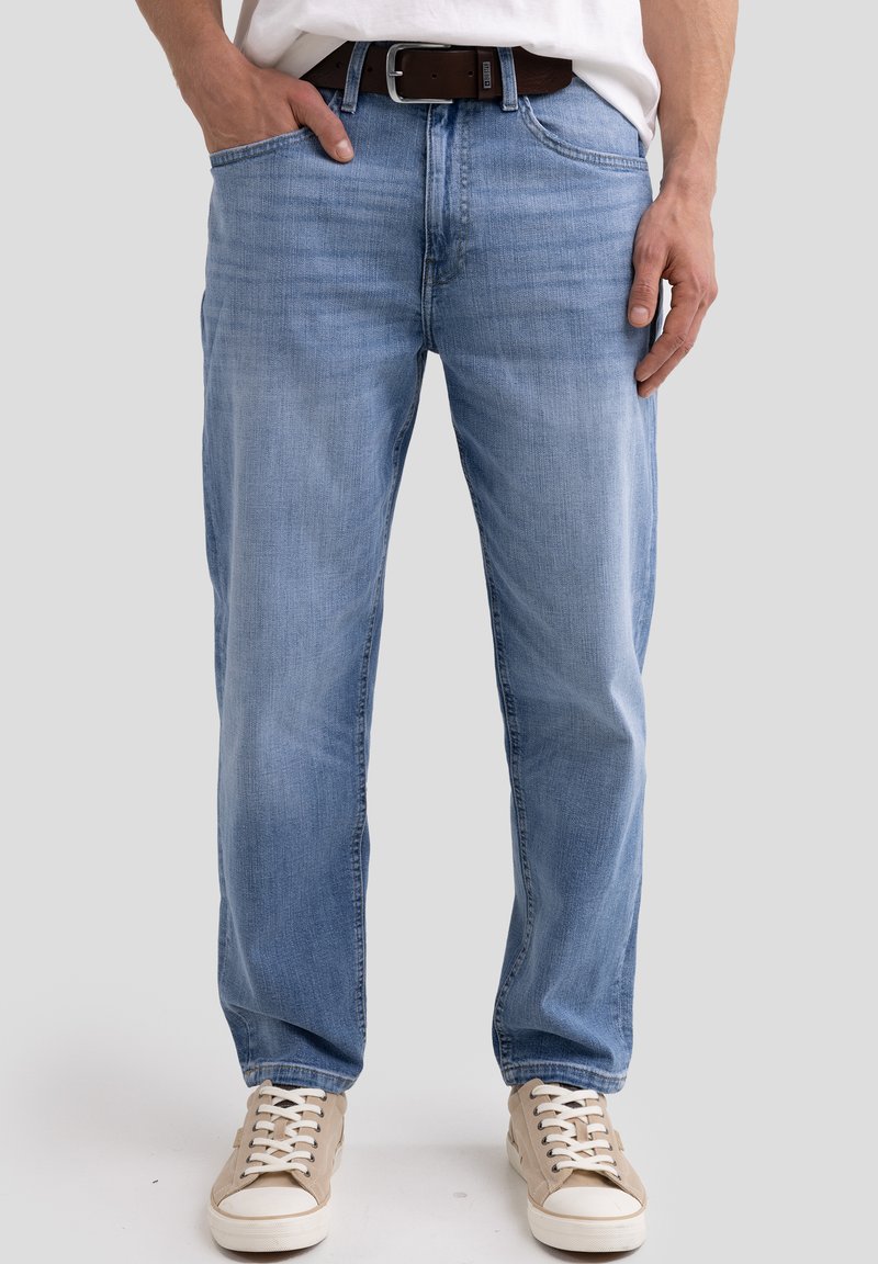 Jean en denim bleu clair avec une coupe décontractée et un design à cinq poches. Ceinture en cuir brun avec une boucle rectangulaire. Baskets beige décontractées.