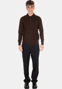 Polo shirt lungo a maniche con lavorazione a maglia color marrone, con colletto e una pattina con tre bottoni, abbinato a pantaloni e scarpe neri.