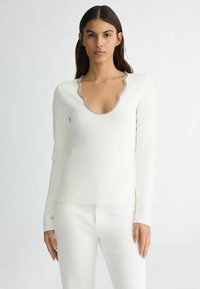 Maglietta bianca a maniche lunghe con scollatura a V profonda e bordo ondulato argentato. Realizzata in un materiale morbido, presenta una superficie liscia e un design aderente.