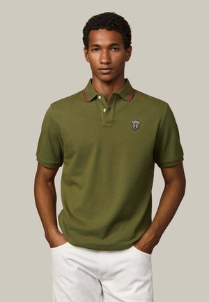 Olivgrünes Polohemd aus Strickstoff, mit einem Kragen mit roten Streifen und einem Logopatch auf der Brust. Kurzärmlig und mit lockerem Schnitt.