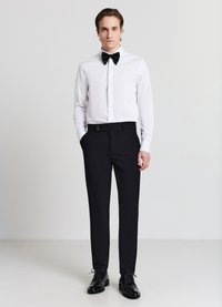 Abito formale composto da una camicia bianca con bottoni, papillon nero e pantaloni neri su misura con dettagli a bottoni. Scarpe eleganti nere lucide.