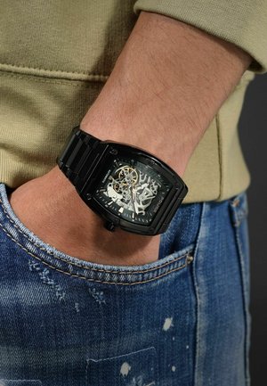 Czarny szkieletowy zegarek z prostokątną kopertą, metalicznymi akcentami i wzorzystą tarczą, noszony na nadgarstku. Tło stanowi dżinsowa tkanina.