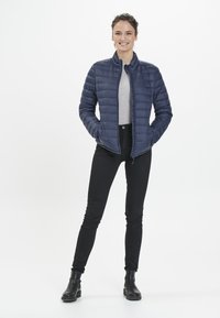 Marinblå puffjacka med hög krage, dragkedjestängning och horisontell quiltad mönster. Bärs över en grå skjorta; matchas med svarta jeans och stövlar.
