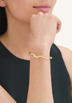 Donna che indossa un elegante braccialetto d'oro con un design ondulato e un piccolo diamante, appoggiando il mento sul pugno su uno sfondo neutro.