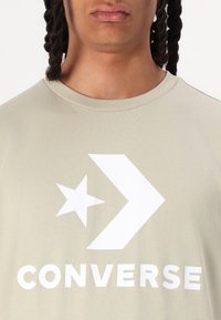 Gai beigā T-kreklu ar apaļo apkakli, kurā ir balts zvaigznes un bultas grafiks līdzās vārdiem "CONVERSE" treknrakstā baltā krāsā.