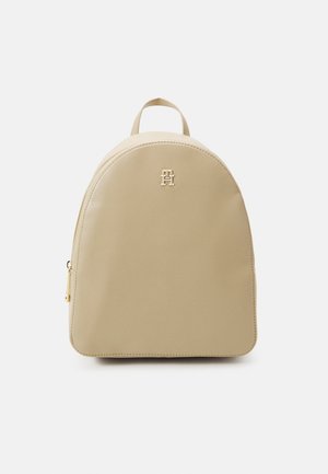 Beige læderrygsæk med afrundet top, lille håndtag, gylden lynlås i siden og et gyldent emblem på forsiden i midten.