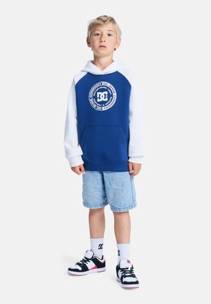 Niebiesko-biała bluza z kapturem z logo, denimowe szorty oraz czarno-białe trampki. Bluza ma kieszeń kangurka i jest zakładana przez głowę.