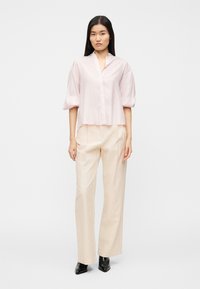 Ανοιχτό ροζ μπλούζα με γιακά και κοντά φουσκωτά μανίκια, με κούμπωμα κουμπιών στο μπροστινό μέρος. Συνδυασμένη με μπεζ πλισέ παντελόνες και μαύρες μπότες με τακούνι.