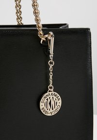 Sac à main en cuir noir texturé avec une bandoulière chaîne dorée et un pendentif rond avec le logo doré DKNY suspendu à la fermeture.