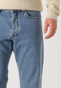 Pantalones vaqueros de mezclilla azul claro con corte recto, que presentan un diseño clásico de cinco bolsillos y una textura desteñida. Mano parcialmente visible a la derecha.
