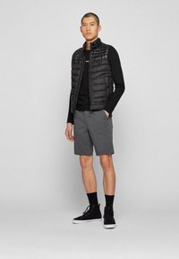 Svart puffervest, ribbet svart langermet skjorte, grå shorts, svarte høye sneakers; stående posisjon, nøytral bakgrunn.