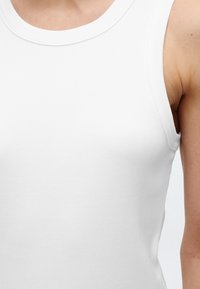 Γυναίκα που φοράει λευκό ριμπαριστό αμάνικο τοπ, σε κοντινό πλάνο στο στήθος και την περιοχή των ώμων, με απλό φόντο.