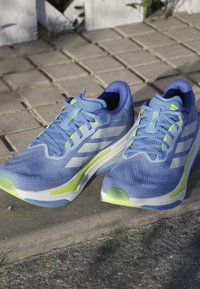 Blue athletic shoes with mesh upper, green accents, and light sole. Features textured surface and reflective details on the sides.