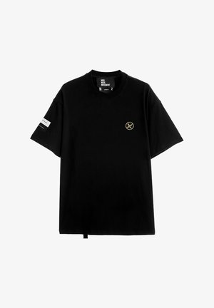 Camiseta de algodón negra con cuello redondo, mangas cortas, emblema dorado en el lado izquierdo del pecho y etiqueta distintiva en la manga.