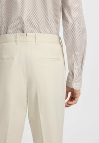 Vaaleanbeiget housut, joissa on sileä pinta ja kaksi takataskua. Paidassa on pehmeissä väreissä oleva geometrinen kuvio ja napilliset hihansuut.