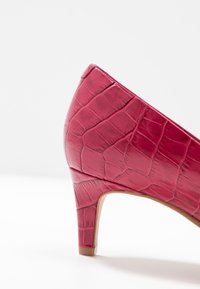 Chaussure à talon haut rose avec une finition texturée ressemblant à de la peau de crocodile. Elle présente un bout pointu et un talon fin. Les bords sont lisses.