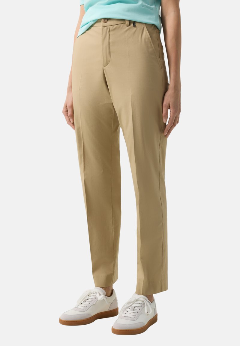 Person wearing beige tapered trousers, light blue top, and white sneakers with brown soles, standing against a plain background.