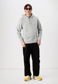 Grå hettegenser med kengurulomme og logo-lapp, kombinert med svarte bukser og gule joggesko. Glatt stoff og avslappet passform.