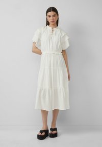 Witte midi-jurk in lagen met een geraffineerde halslijn en korte mouwen, een getailleerde taille en een textuurstof. Zwarte plateau-schoenen.