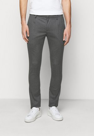 Uomo che indossa pantaloni grigi slim fit e sneakers bianche, in piedi contro uno sfondo chiaro e uniforme.